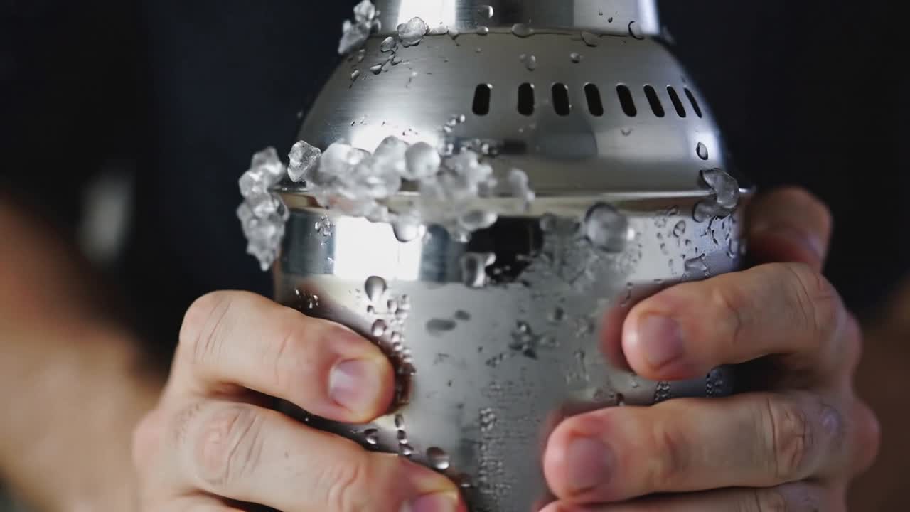Mains de barman saisissant un shaker, déplaçant la glace et projetant des gouttelettes pour rafraîchir un cocktail.