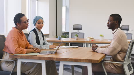 an woman and a muslim woman co workers interview a young man sitting at a table in the office 15