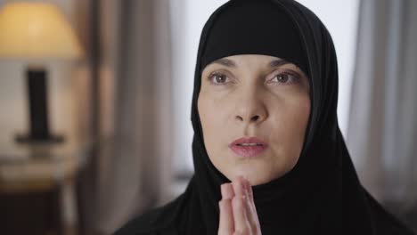 close-up face of young beautiful muslim woman holding hands together and praying. lady in black traditional hijab talking with god. culture, religion. camera moving around from right to left.