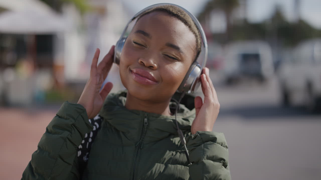 retrato de una joven mujer afroamericana con auriculares escuchando música bailando feliz disfrutando de cálidas y vibrantes vacaciones de verano en el fondo de la playa urbana