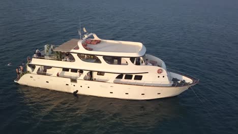 Orbit-shot-Of-White-Yacht-Anchored-In-Middle-Of-Blue-Ocean-While-People-Swim-Around,-Cyprus