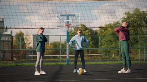 three athletes standing on outdoor volleyball court, working out by holding their heads and waist, volleyball rests in front, with sports arena and residential building in background