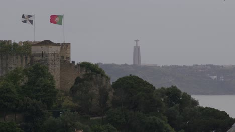 Statische-Aufnahme-Der-Portugiesischen-Flagge,-Die-An-Einem-Windigen-Tag-Mit-Einer-Jesusstatue-Im-Hintergrund-In-Lissabon,-Portugal,-Weht