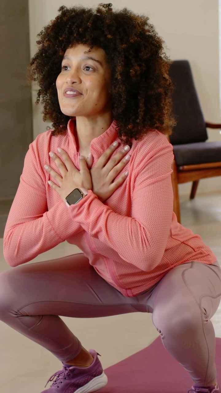 Vídeo vertical: Mulher respirando com top coral fazendo agachamento guiado em casa, em tapete roxo, com relógio