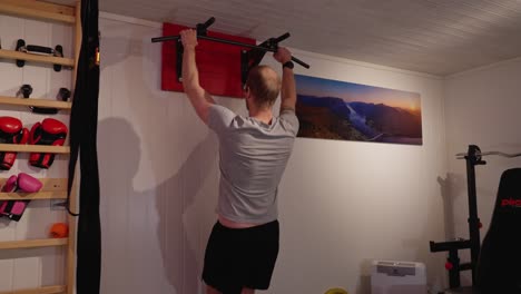 man in late thirties performs pull ups on a bar in his home gym basement with rib wall and weight bench visible on sides