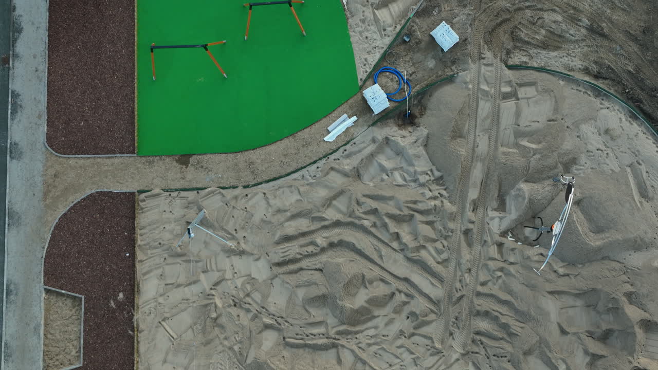 Aerial view of a construction site with a green turf area and various materials on sand, illustrating ongoing landscape development