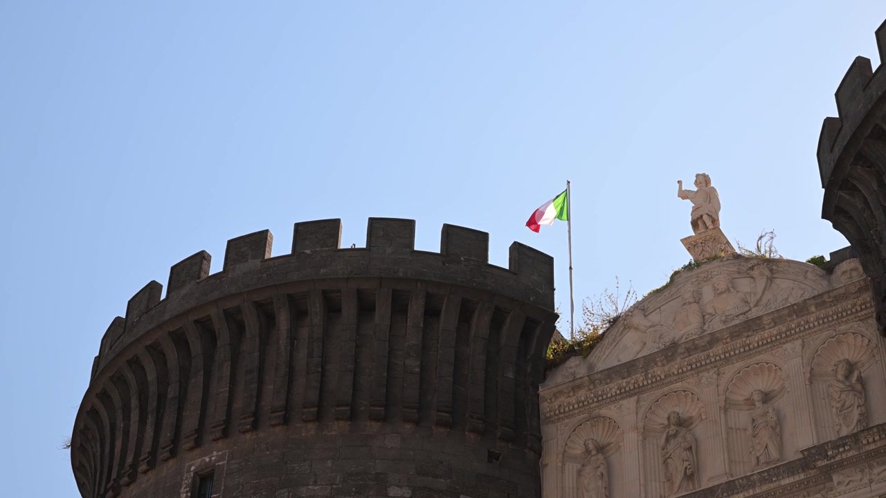 La bandiera italiana sventola nella brezza sopra la torre in pietra e le statue di Castel Nuovo contro un cielo azzurro limpido