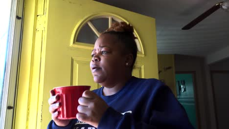 medium shot of african american gen z female drinking a hot drink from a red mug while looking out the front door of her home