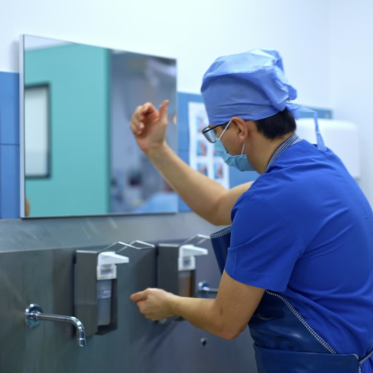 Surgeon in uniform, mask, cap, glasses and apron comes up to a big metal sink. Doctor presses on dispenser with his elbow and spreads antiseptic over his arms