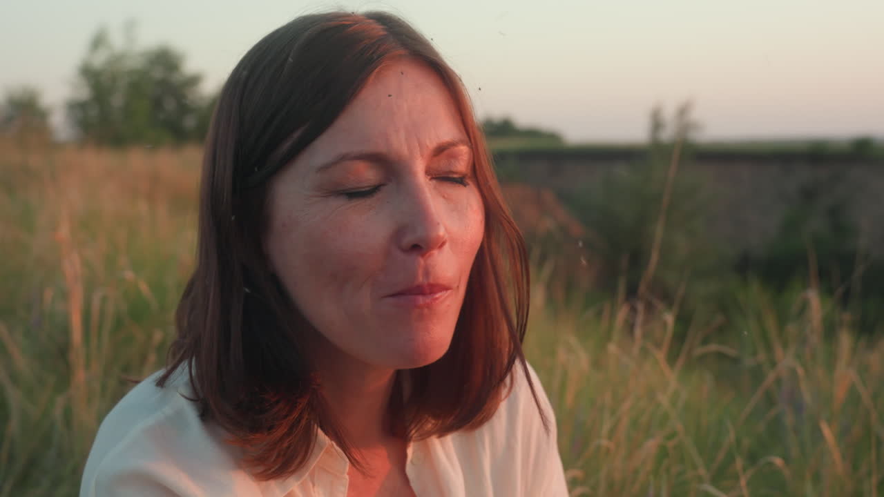 Elegant woman seated in tall grass holding ripe plum near mouth, slowly enjoying sweet fruit in warm golden hour light, with serene countryside background