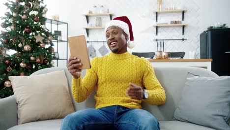 Happy-Handsome-Male-In-Xmas-Hat-Videochatting-On-Tablet-While-Sitting-Near-Decorated-New-Year-Tree-At-Home