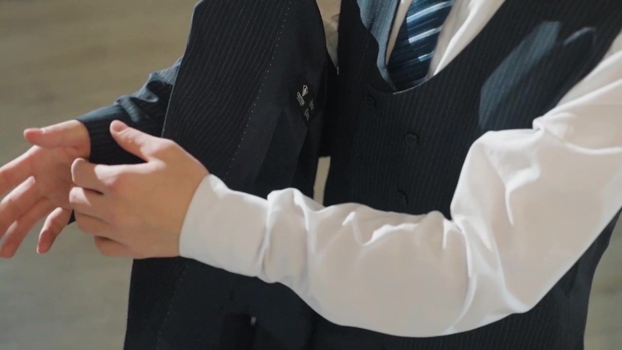 Giovane uomo asiatico che indossa un gilet in una stanza illuminata delicatamente, sistema la cravatta a righe e i bottoni, primo piano delle mani che lisciano il tessuto di lana, stile uniforme scolastica, tranquilla attesa e preparazione attenta.