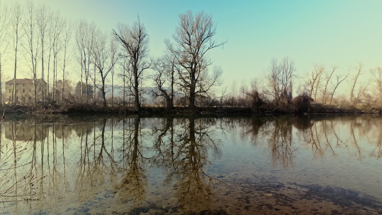 Winter leafless trees along riverbank reflect in slow flowing water