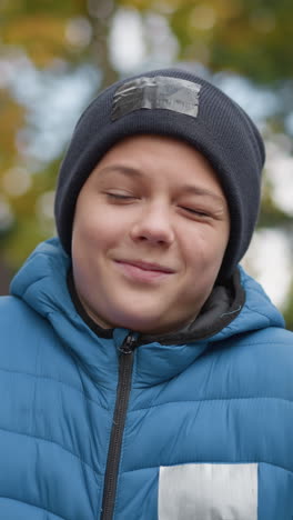 niño con gorra negra y chaqueta azul de pie al aire libre, sonriendo y disfrutando del momento con una expresión feliz, vegetación borrosa y paisaje de otoño en el fondo