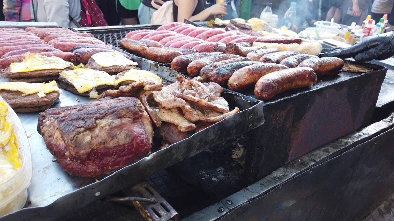 Close-up de carne grelhada argentina e linguiças tradicionais em um mercado de asado de rua, com Choripan e hambúrgueres com queijo.