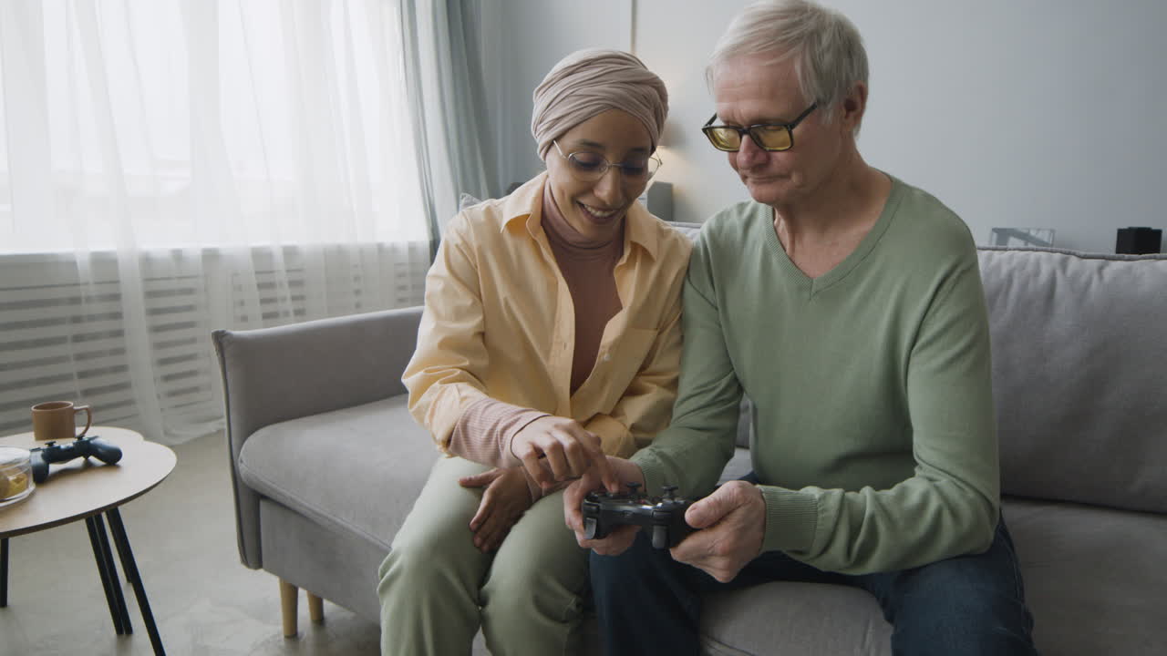 Middle Aged Arabic Woman Explaining To A Senior Man How To Use The Game Controller While Sitting Together On Couch At Home