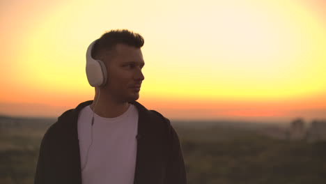 smiling and laughing handsome young man listening music from his smartphone in wireless headphones. in slow motion a man stands on the roof at sunset and looks at the beautiful view from the height of the city