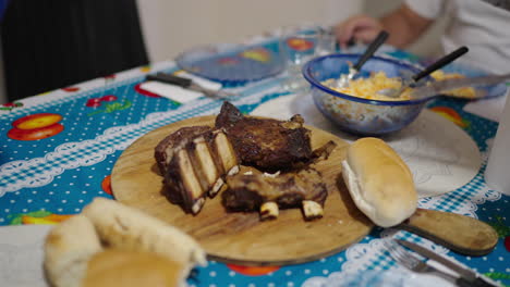 hand slices succulent asado ribs on a wooden board in slow motion, accompanied by bread rolls and a bowl of salad, all set on a vibrant tablecloth in an authentic argentine home, camera slow motion.