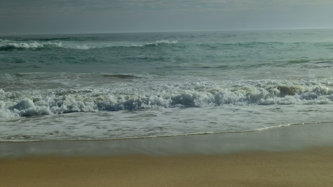 Powerful ocean waves roll onto a sandy shore bank, creating white foam as they crash and retreat, shaping the coastline with rhythmic motion.