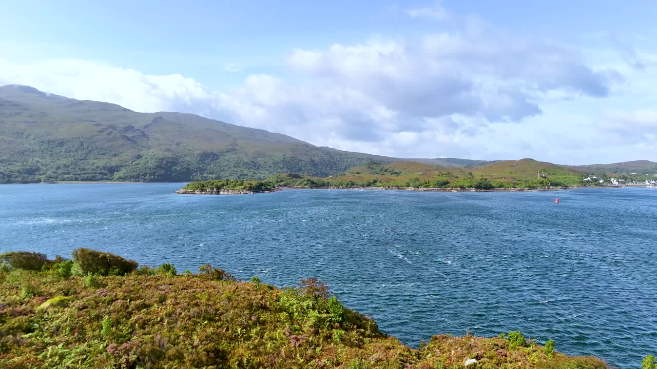 Aerial camera slowly pans over a lush, rocky peninsula toward a blue loch, revealing distant hills under bright daylight in the Scottish Highlands