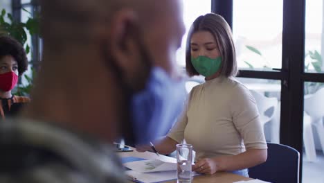 diverse business people wearing face masks sitting using laptops going through paperwork in office