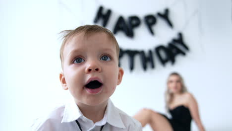 grey-eyed caucasian toddler boy in white shirt and black tie stands in front of camera. blond woman sits at blurred backdrop. happy birthday sign on the wall.