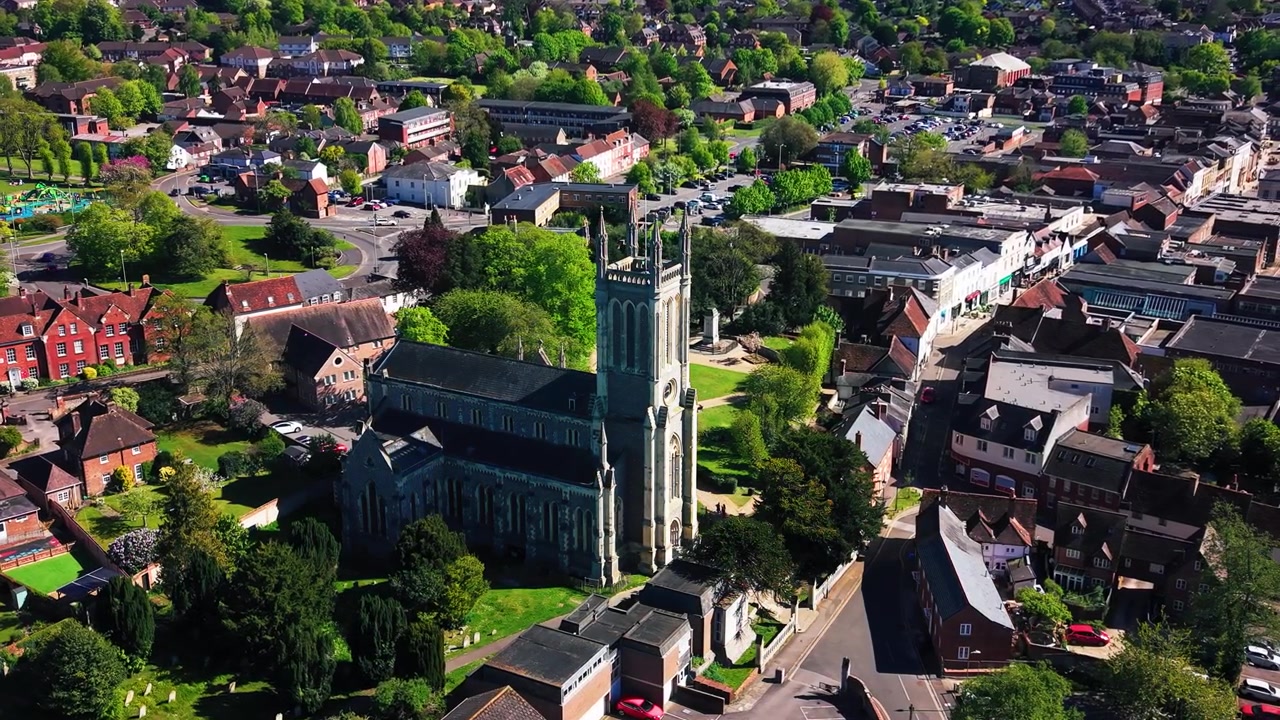 Letecké záběry dronu obíhajícího kostel svaté Marie v Andoveru, ukazující historickou kostelní věž, hřbitov, okolní ulice, střechy, zelené plochy a širší krajinu města Hampshire za slunečného dne