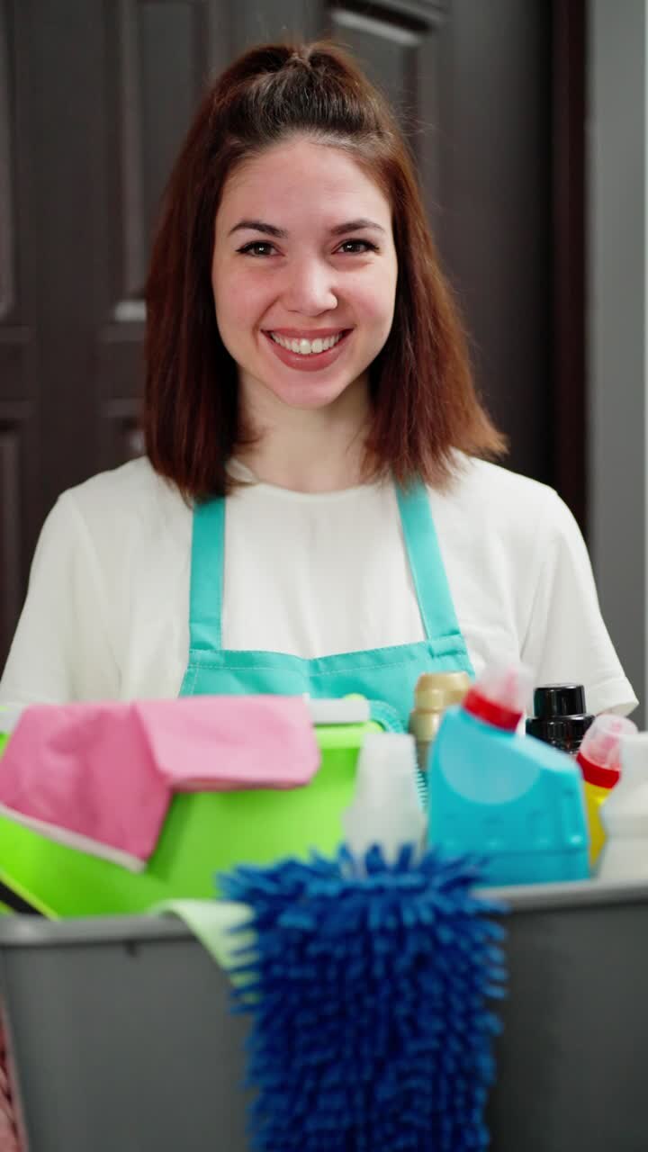 Vertical video portrait of a happy girl professional cleaner in blue aprons with many cleaning items in her hands in the house