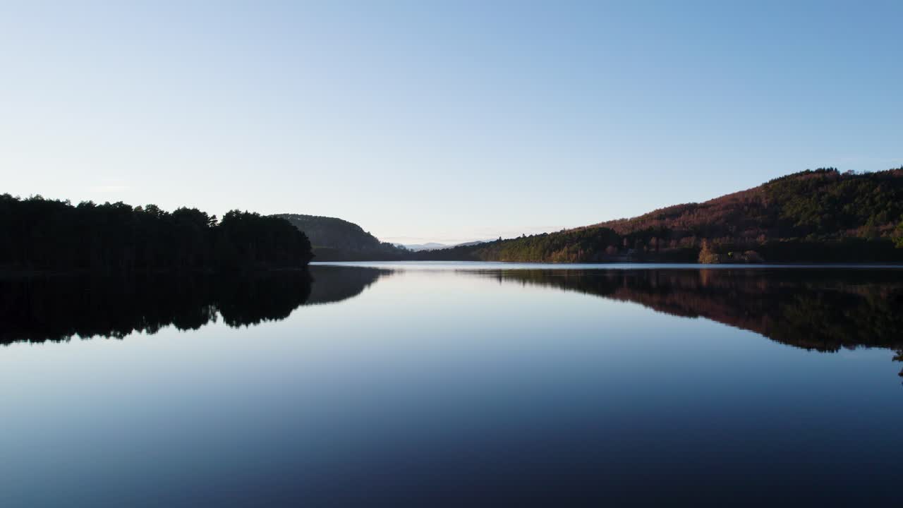imágenes aéreas de drones que se elevan sobre la superficie de aguas tranquilas con reflejos en el parque nacional de cairngorms, escocia, para revelar un bosque nativo y un paisaje montañoso al atardecer