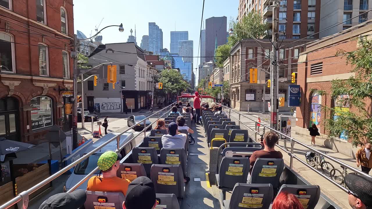 First-person view of Toronto King Street from moving hop-on hop-off sightseeing bus, with tourists seated enjoying tour among towering skyscrapers and bustling car traffic, Canada