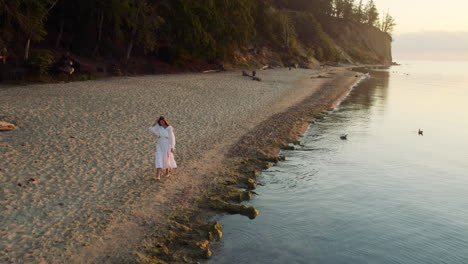 Beautiful-woman-in-white-flowing-dress-plays-dancing-on-tropical-beach-at-sunset