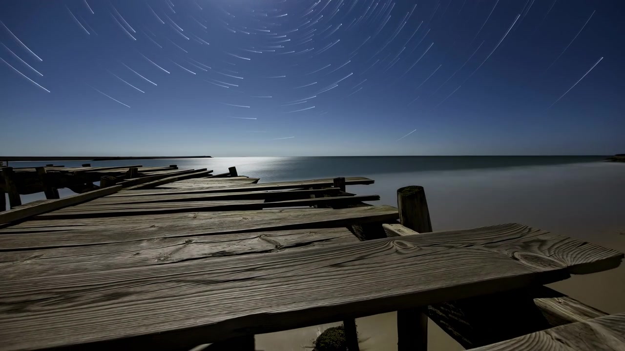 Assi di legno stagionate si estendono dal primo piano verso una riva tranquilla, creando una forte prospettiva sotto un cielo scuro e limpido pieno di stelle.
