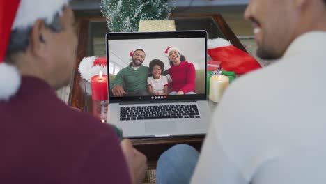 Padre-E-Hijo-Birraciales-Sonrientes-Usando-Una-Computadora-Portátil-Para-Una-Videollamada-Navideña-Con-Una-Familia-Feliz-En-La-Pantalla