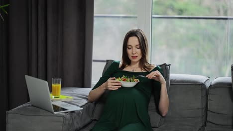 Pregnant-woman-sitting-on-the-sofa-at-home-using-laptop-and-eats-salad