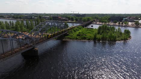 Increíble-Toma-De-Seguimiento-Del-Tren-Cruzando-El-Puente-Ferroviario-Sobre-El-Río-Daugava-En-Riga,-Letonia