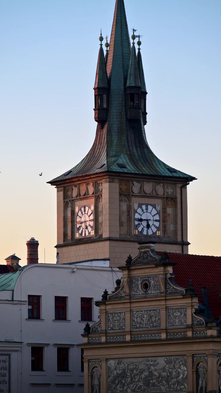 Vista verticale di una torre storica con doppi quadranti e una guglia di rame verde al tramonto, Praga, Repubblica Ceca