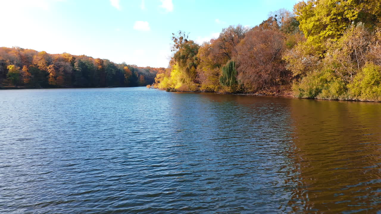 Calm dark surface of river flowing between the banks covered with woods. Bright trees overgrow the waterfront. Sunny autumn day in nature.