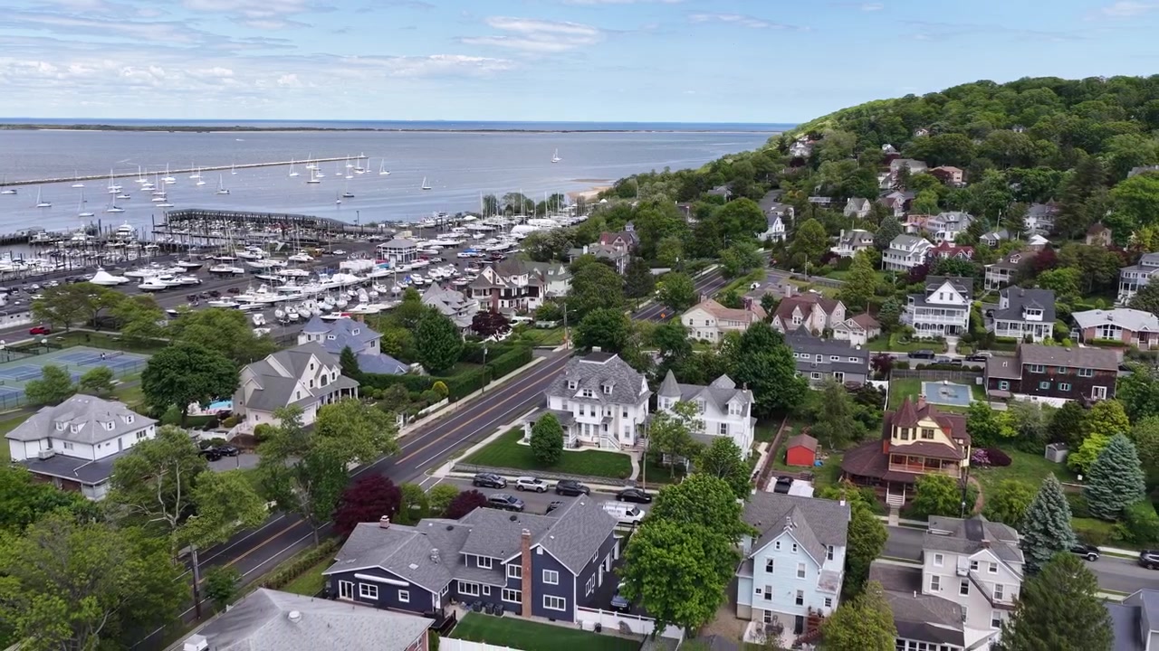 Vista aerea di Atlantic Highlands, NJ che mostra la baia di Sandy Hook e un porto turistico con barche ormeggiate, adiacente a parcheggi e case residenziali su un verde pendio sotto un cielo parzialmente nuvoloso.