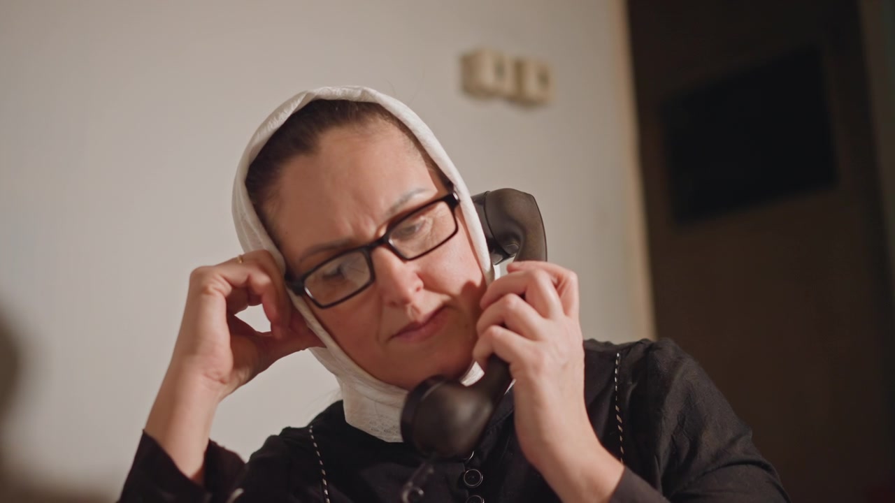 Moden kvinne med hodeskjerf holder telefon, lukker øynene med et mykt utpust, ømt uttrykk, telefonrør med ledning og briller, varmt dempet belysning, filmatisk nærbilde, subtil spenning, privat stue