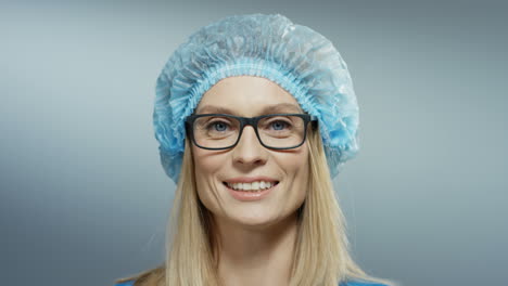 Close-Up-Of-The-Pretty-Female-Young-Doctor-In-Glasses,-Blue-Hat-Smiling-Cheerfully-To-The-Camera