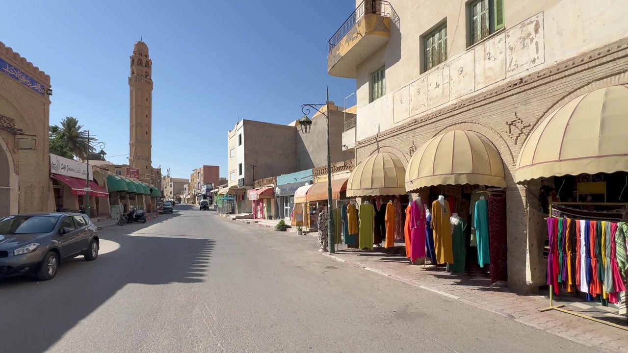 Tunus'un Tozeur şehrinde, sakin ve güneşli bir cadde boyunca sıralanmış geleneksel giysi tezgahları, uzakta yüksek bir cami minaresi ve seyrek geçen arabalar.