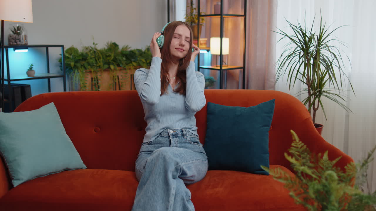 무선 헤드폰을 입은 행복하고 편안한 소녀가 춤을 추고 집 소파에서 좋아하는 록 앤 롤 음악을 듣고 있습니다.