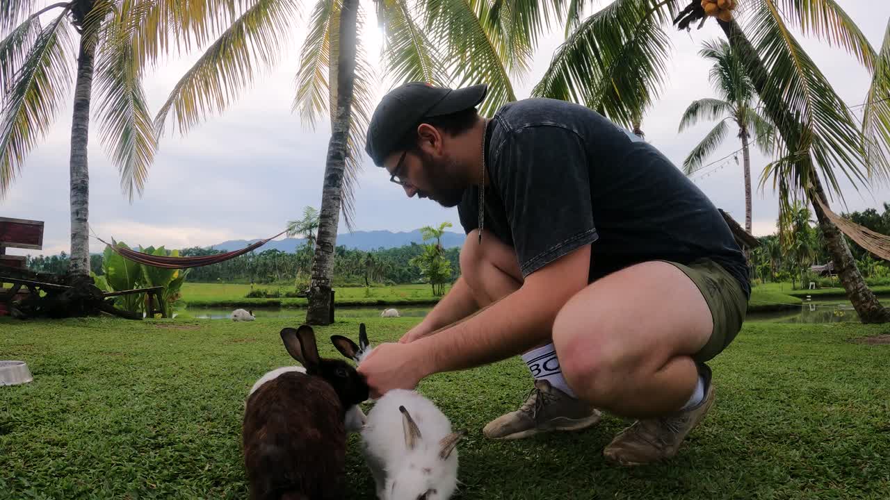 Side Portrait Of A Cheerful Male Tourist Petting Herd Of Rabbits On The Grass In A Nature Park In The Philippines. full shot