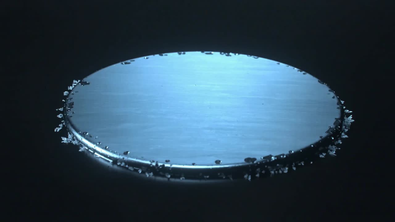 Dendritic crystals growing from rim to center on metal disk in dark studio, droplets seeding growth