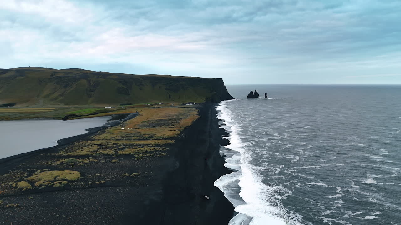 Black narrow sandy beach line stretching to the steep mountain. Few sharp rocks stick out from waterscape. Shore of Iceland on a grey dull day.