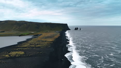 black narrow sandy beach line stretching to the steep mountain. few sharp rocks stick out from waterscape. shore of iceland on a grey dull day.