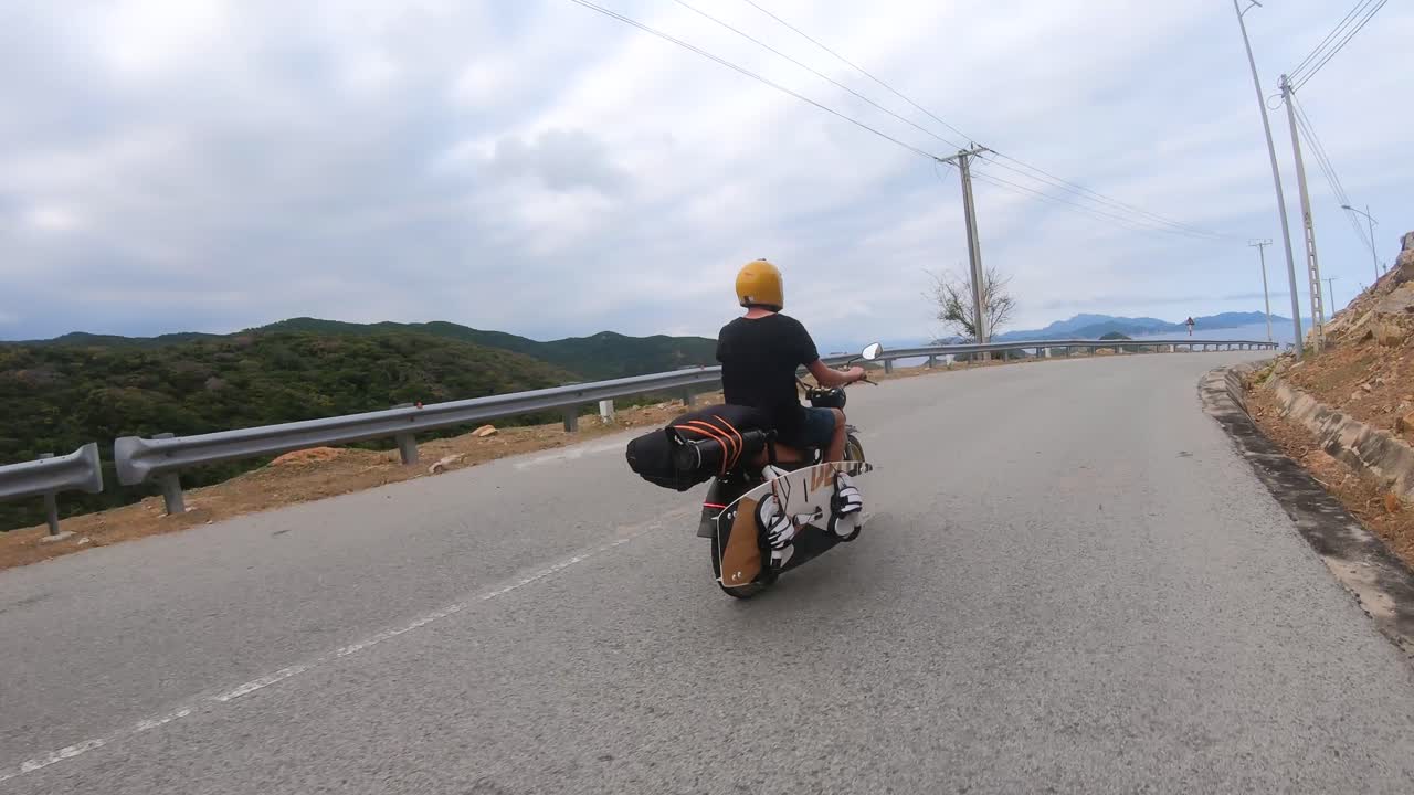 Vista posteriore di un motociclista su strada costiera con una tavola da kitesurf legata alla moto.