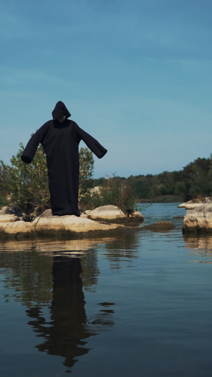 Evil witch in black cloak with hood on stones near the river. Divination by the water. Halloween costume. Vertical video