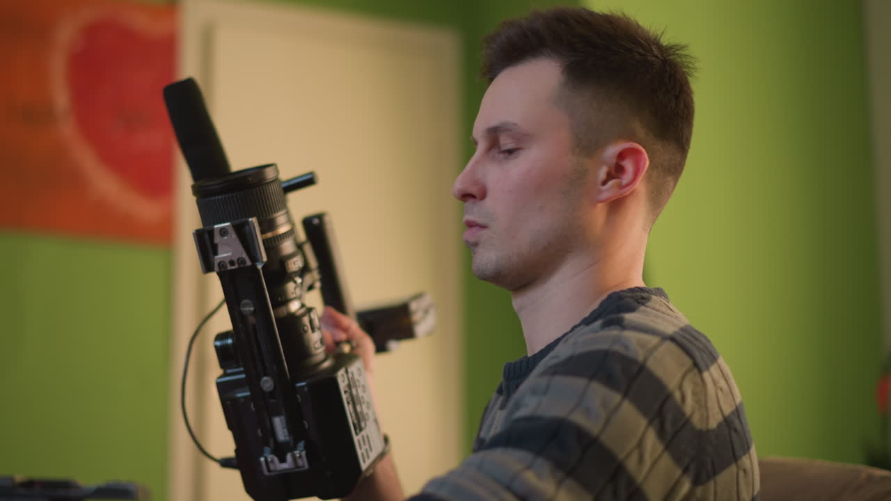TV technician focusing on camera setup. Technician holds camera rig with microphone and lens, adjusting equipment with serious concentration in indoor studio with green wall