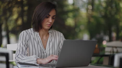 Retrato-Mujer-Seria-Morena-Grupo-étnico-Hispano-árabe-Se-Sienta-En-Una-Mesa-En-Un-Café-De-Verano-Con-Una-Computadora-Portátil.-Escribe-En-El-Teclado-Y-Recibe-Capacitación-En-Línea-Compra-Y-Negocia-Con-Un-Banco-De-Internet